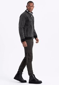 Harmaa pommittajatakki mustilla hihansuilla ja edessä vetoketju, yhdistettynä oliivinvihreisiin cargo-housuihin ja mustiin nauhattomiin saappaisiin. Hieno kangas, minimalistinen muotoilu.