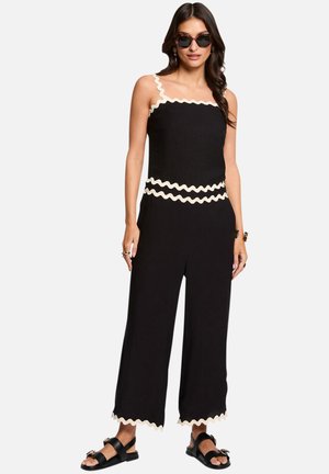 Femme portant une combinaison noire sans manches avec un bord ondulé blanc, des sandales noires, des lunettes de soleil et des bijoux minimalistes, debout devant un fond blanc.