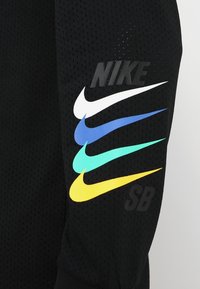 Svart kortärmad tröja med meshtextur, med en stor "Nike"-logotyp och fem färgade swooshes: vit, blå, grön, ljusblå och gul.