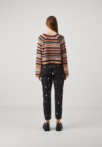 Femme aux cheveux longs et raides portant un pull multicolore à rayures, un jean noir avec des motifs de fleurs blanches et des chaussures noires cloutées, debout immobile.