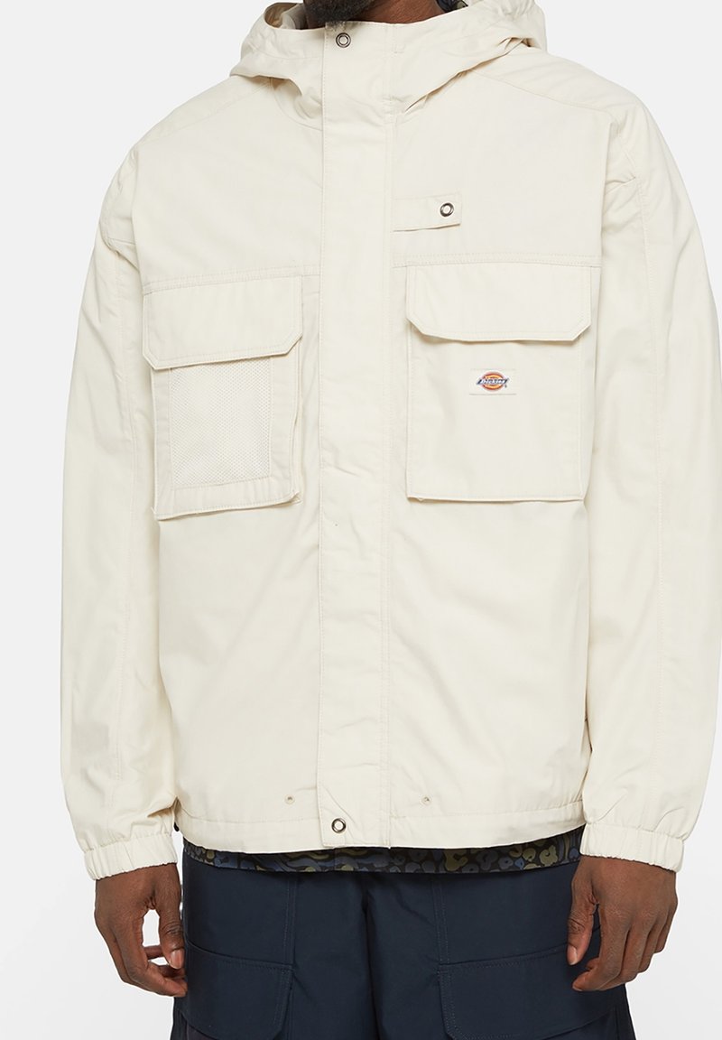 Světlý béžový kabát s kapucí, dvěma velkými předními kapsami, elastickými manžetami a detailem síťované kapsy. Obsahuje logo nášivku. Hladká textury tkaniny.