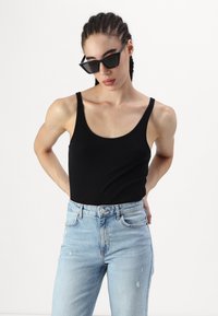 Crna majica bez rukava na modelu, u kombinaciji s iznošenim svijetloplavim trapericama. Majica ima zaobljeni izrez i usku siluetu.