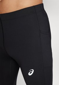 Sorte atletiske leggins lavet af strækbart materiale, med flade sømme, en sidelomme og et hvidt logo på venstre lår.