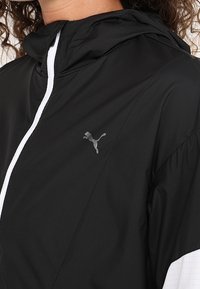 Lekka czarna kurtka wiatrówka z kapturem, kontrastowymi białymi detalami i subtelnym szarym logo Puma. Teksturowany materiał. Zapięcie na zamek błyskawiczny.