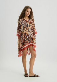 Kaftan in stampa animale trasparente con scollo a V, in tonalità arancioni e beige, caratterizzato da maniche e orlo bordati di rosso. Abbinato a sandali neri.