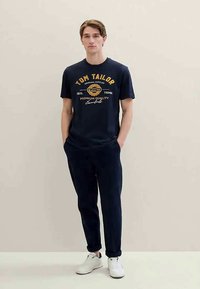 Ναυτικό μπλουζάκι με κίτρινο γραφικό κείμενο και λογότυπο, συνδυασμένο με σκούρες φόρμες και λευκά αθλητικά παπούτσια. Το μοντέλο στέκεται σε ένα ανοιχτό φόντο.