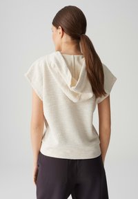 Light beige short-sleeve top with a hood, featuring a textured fabric and a relaxed fit, paired with dark pants.