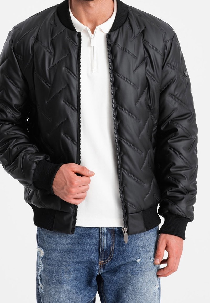 Homme portant une veste bomber matelassée noire sur une chemise blanche zippée et un jean bleu déchiré, tenant la fermeture éclair de la veste près de la poitrine.
