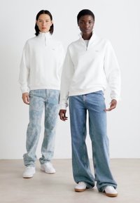 Dos personas llevan sudaderas blancas con cremallera. Una tiene pantalones de patrón azul claro, y la otra lleva jeans azules clásicos y zapatillas blancas.