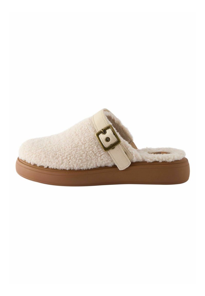 Chausson en crêpe de couleur crème floue avec une texture douce, présentant une sangle légère contrastante et un détail de boucle, ainsi qu'une semelle en caoutchouc marron.
