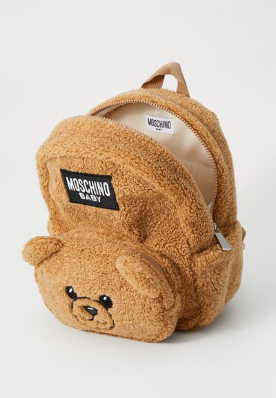 Sac à dos en forme d'ours en peluche beige, avec une texture douce, un patch logo noir et une fermeture éclair. Comprend des oreilles arrondies et des détails faciaux brodés.