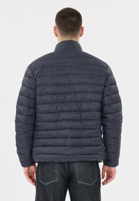 Marinblå puffjacka med horisontell quiltad mönstring, hög krage och elastiska ärmslut, sedd från baksidan. Slät, glänsande tygtextur.