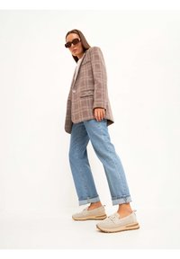 Blazer à carreaux marron, chemise grise claire, jeans bleu droit et baskets perforées beiges avec des semelles et des lacets marron, le tout agrémenté de lunettes de soleil noires.