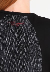 Schwarzes Oberteil mit einem strukturierten Rückenteil in einem Fischgrätenmuster in Schwarz und Weiß, verziert mit dezenten roten Stickereien.