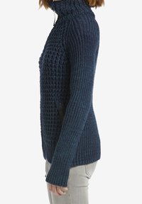 Tmavě modrý pletený svetr s vysokým límcem, zdobený strukturovaným žebrováním a boční kapsou se zipem. Dlouhé rukávy přesahují zápěstí.