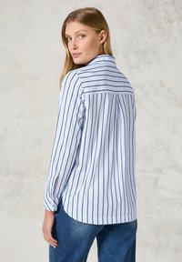 Light blue long-sleeve shirt featuring vertical black stripes, a collar, and buttoned cuffs. The design has a relaxed fit with a back pleat.