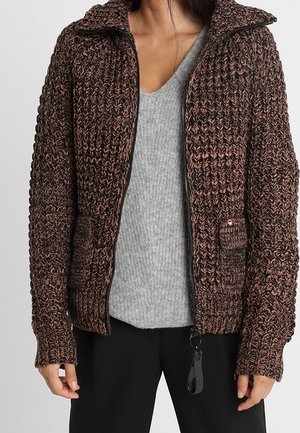 Brun strikket jakke med et tekstureret mønster, lynlås lukning, to forlommer og krave. Bæret over en grå sweater.