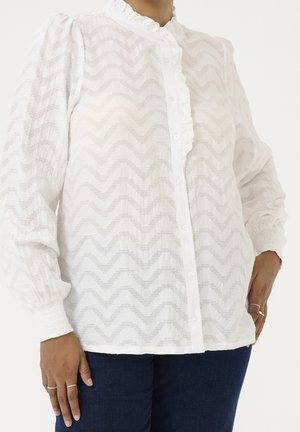 Biała bluzka z długim rękawem z subtelnym wzorem zygzaka, zapinana na guziki z przodu, z falbankowym kołnierzem i marszczonymi mankietami, noszona z ciemnoniebieskimi dżinsami.