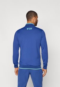Plava jakna na zip napravljena od glatkog materijala, sa rebrastim ovratnikom i manžetama. Svijetloplavi detalji na ovratniku i donjem rubu. Logo na leđima.