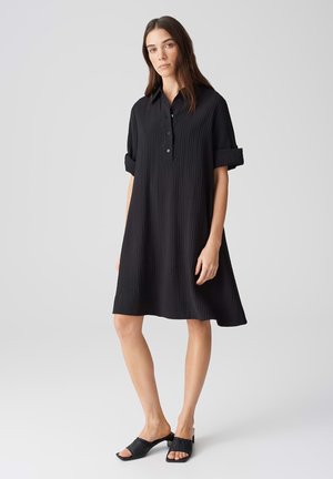 Schwarzes, knielanges Hemdkleid mit strukturierter Rippoptik, Knopfkragen und umgeschlagenen Kurzärmeln. Kombiniert mit schwarzen Sandalen mit offenem Zehen.