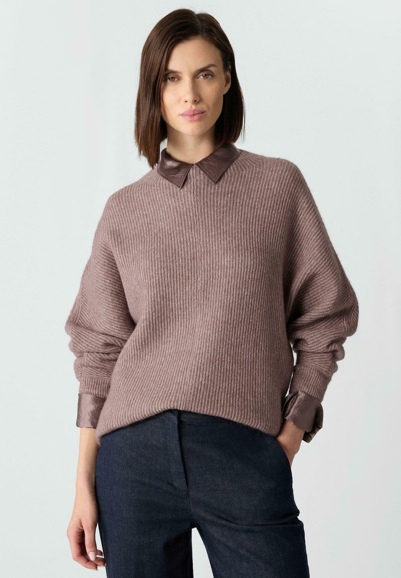 Jasnobrązowy prążkowany sweter z brązowym skórzanym kołnierzem, opuszczonymi ramionami i szerokimi rękawami, zestawiony z ciemnoniebieskimi spodniami.