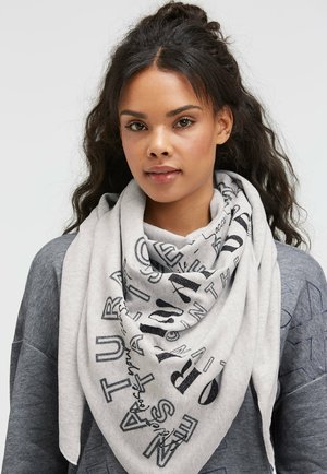 Young woman with long dark hair wearing a gray scarf with black printed text over a gray sweatshirt against a plain background.