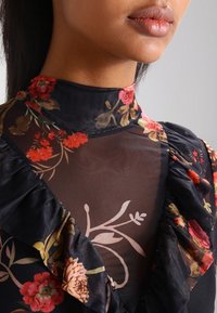 Blusa nera a fiori con collo alto, tessuto trasparente, accenti arricciati e motivi floreali colorati in tonalità di rosso, rosa e verde.