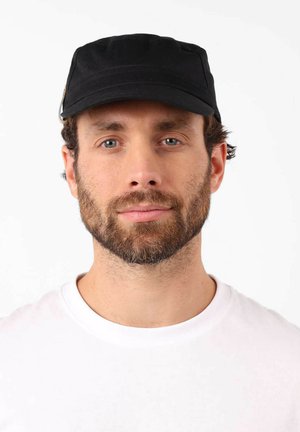 Homme barbu à la peau claire et aux yeux bleus, portant une casquette noire et une chemise blanche, faisant face à la caméra avec une expression neutre sur un fond blanc.