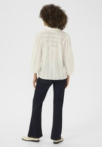 Blusa bianca a righe con una vestibilità rilassata, maniche a palloncino e un dettaglio sullo yoke posteriore, abbinata a jeans in denim scuro a zampa d'elefante e scarpe color crema.