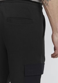 Black cotton sweatpants feature a side cargo pocket with a navy flap, elastic waistband, and smooth texture, shown from the back.