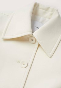 Crème-kleurige blouse met een gestructureerde kraag, twee witte knopen en een gladde stofstructuur met subtiele glans. Merkenlabel zichtbaar aan de binnenkant.