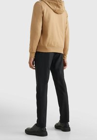 Hoodie in tan stof met geribde manchetten, gecombineerd met zwarte tapered pants en zwarte schoenen. Minimalistisch ontwerp, geen zichtbare logo's.