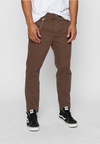 Brune denimjeans med slim fit, med en metallkjede og glidelås i hemmet. Kombinert med svarte sneakers og en hvit T-skjorte.