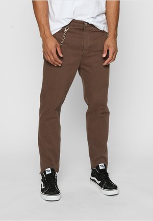 Jeans in denim marroni con una vestibilità slim, dotati di una catena in metallo e di un orlo con zip. Abbinati a sneaker nere e a una t-shirt bianca.