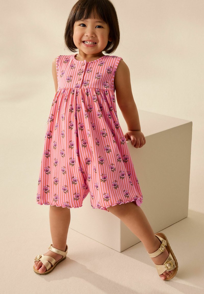 Giovane ragazza sorridente che indossa un vestito rosa floreale senza maniche e sandali beige, in piedi con una mano su un blocco bianco.