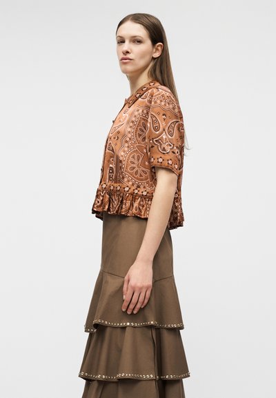 Νεαρή γυναίκα που φοράει καφέ κοντομάνικη μπλούζα με σχέδια και καφέ φούστα πολλαπλών στρωμάτων με διακοσμητική τρέσα με τρουκς, στέκεται πλευρικά μπροστά σε απλό φόντο.