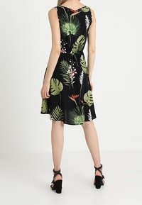 Svart ärmlös klänning med figurnära midja och utsvängd kjol, prydd med gröna blad- och blommönster. Bärs med svarta klacksandaler.