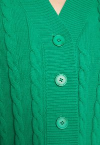 Cardigan en maille verte avec un col en V, des détails en maille torsadée sur les côtés, et trois gros boutons verts sur le devant.