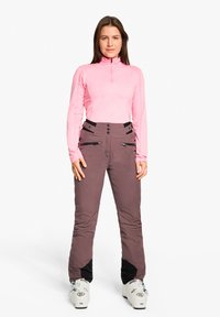 Hellrosa Langarm-Zipper-Top kombiniert mit braunen Skihosen mit schwarzen Akzenten, Reißverschlusstaschen und verstellbarem Bund. Die Skischuhe sind weiß.
