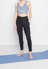 Brassière de sport bleu à motifs avec détail découpé, associée à un pantalon de sport noir fuselé avec poches latérales, posés sur un tapis de yoga bleu.