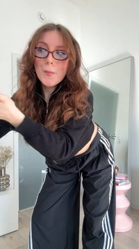 Giacca nera con zip e pantaloni sportivi neri larghi con strisce bianche. Occhiali tartarugati. Capelli mossi e castani. Ambiente interno luminoso.
