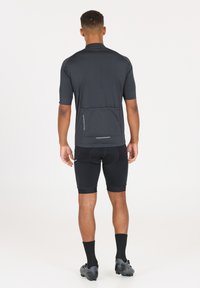 Schwarzes Sporttrikot mit kurzen Ärmeln, verfügt über ein Design mit einer Rückentasche. Kombiniert mit schwarzen Radlerhosen und dunklem Schuhwerk.