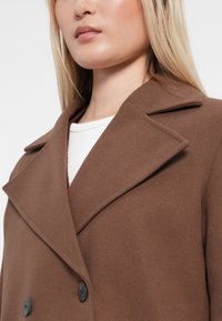 Brun ullblandingsjakke med dobbelbrystet design, store reverser og svarte knapper; glatt tekstur, kne-lengde silhuett.