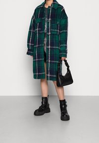 Grön och marinblå rutigt överdimensionerad kappa med två framfickor, buren över en grå topp, ihop med svarta kraftiga stövlar och en liten svart väska.