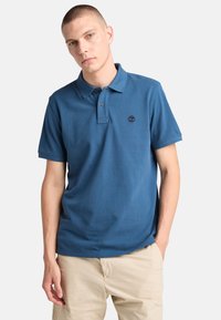 Blå pikétröja i bomull, med krage, tvåknappsslå och en liten broderad logotyp på bröstet. Korta ärmar.