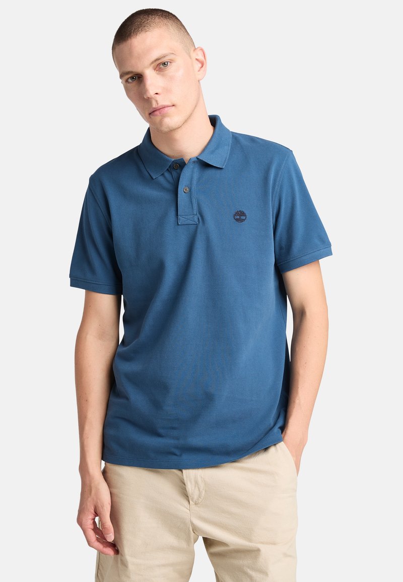 Blå pikétröja i bomull, med krage, tvåknappsslå och en liten broderad logotyp på bröstet. Korta ärmar.