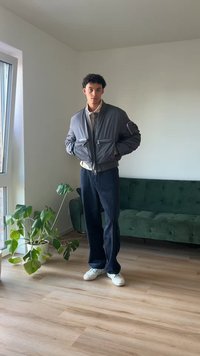 Hall värvi bomber-jakk, millel on lukuga taskud, beež särk, tumedad teksapüksid ja valged spordijalatsid, eksklusiivselt hästi valgustatud ruumis, kus on roheline diivan ja taim.