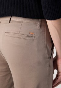 Pantalons beige avec une texture lisse, comprenant une poche arrière boutonnée et une petite étiquette orange "BOSS". Le tissu semble léger.