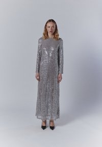 Sidabrinė blizgučiais puošta suknelė ilgomis rankovėmis su aukštu apykakle ir sudėtingais raštais; modelis dėvi juodus smailianosčius batus prieš šviesų foną.