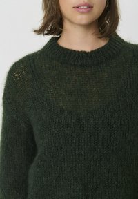 Dunkelgrüner Strickpullover mit rundem Ausschnitt, gefertigt aus lockerem, strukturiertem Garn und einem leicht transparenten Bereich auf der Brust.